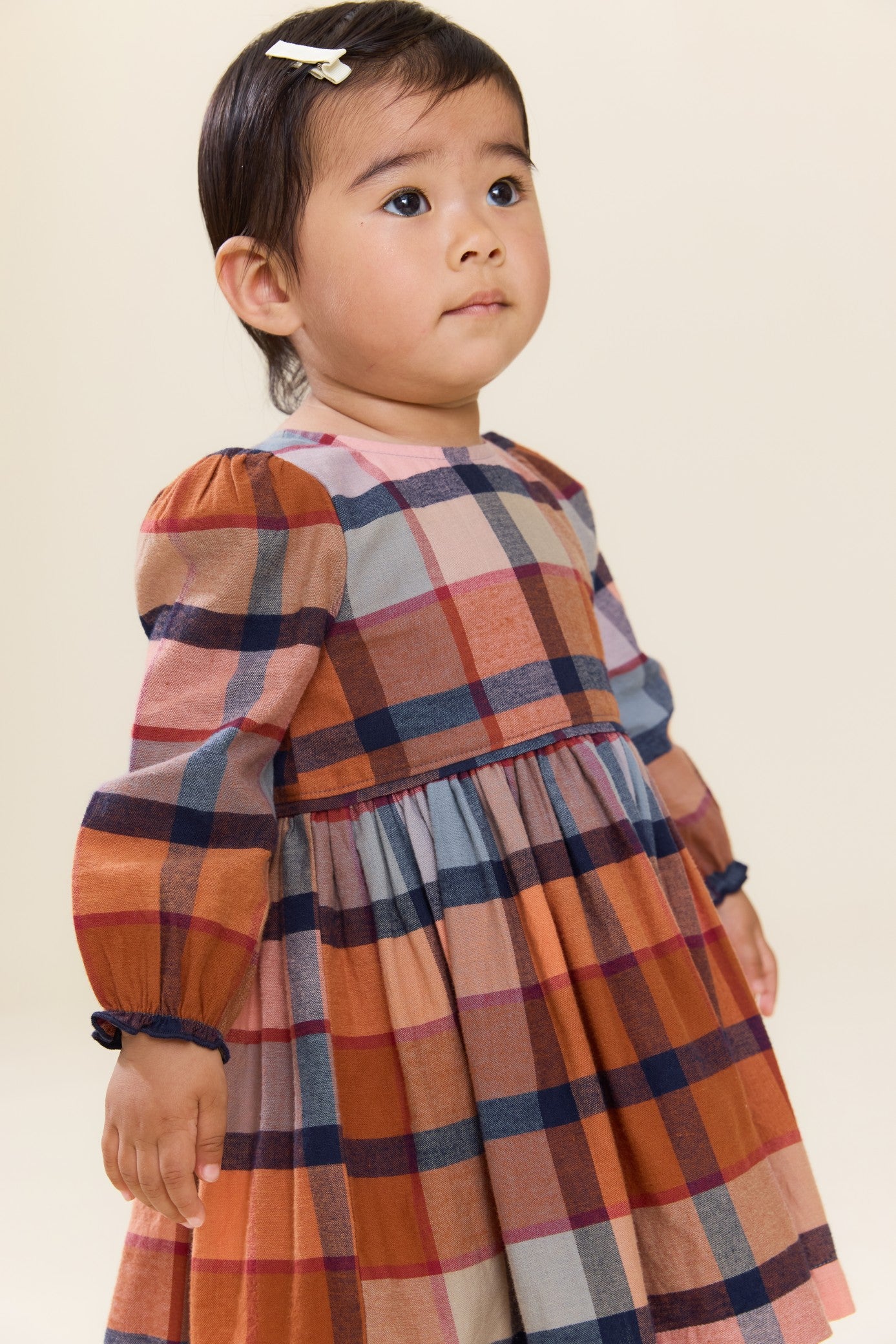 Young child wearing a Pink Chicken long sleeve plaid dress in warm rust blue and cream tones with a gathered skirt and hair clip posing indoors