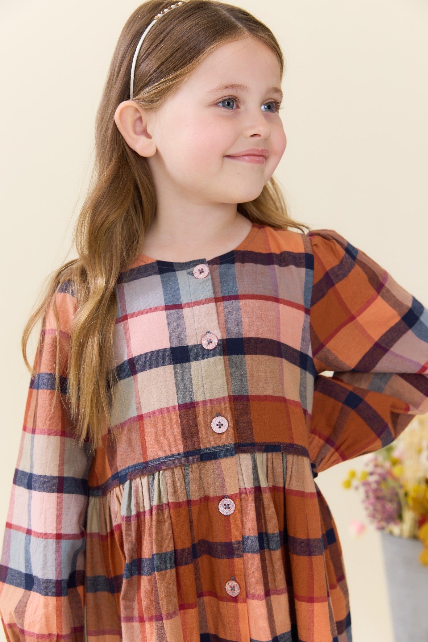 Young girl wearing a warm brown and navy plaid dress with button details and a gathered skirt from Pink Chicken