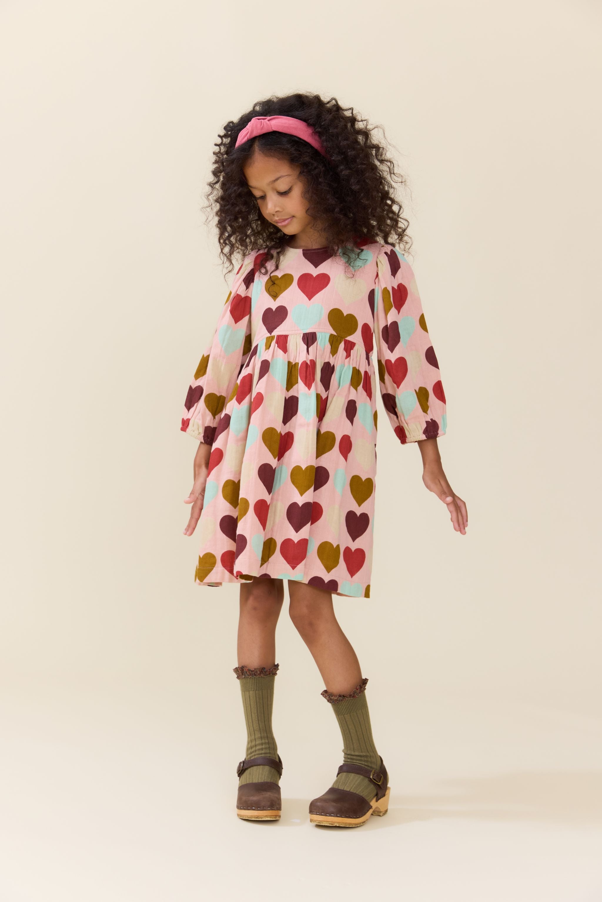 Young girl wearing a long sleeve cream dress covered in colorful hearts with a pink headband olive socks and brown shoes from Pink Chicken