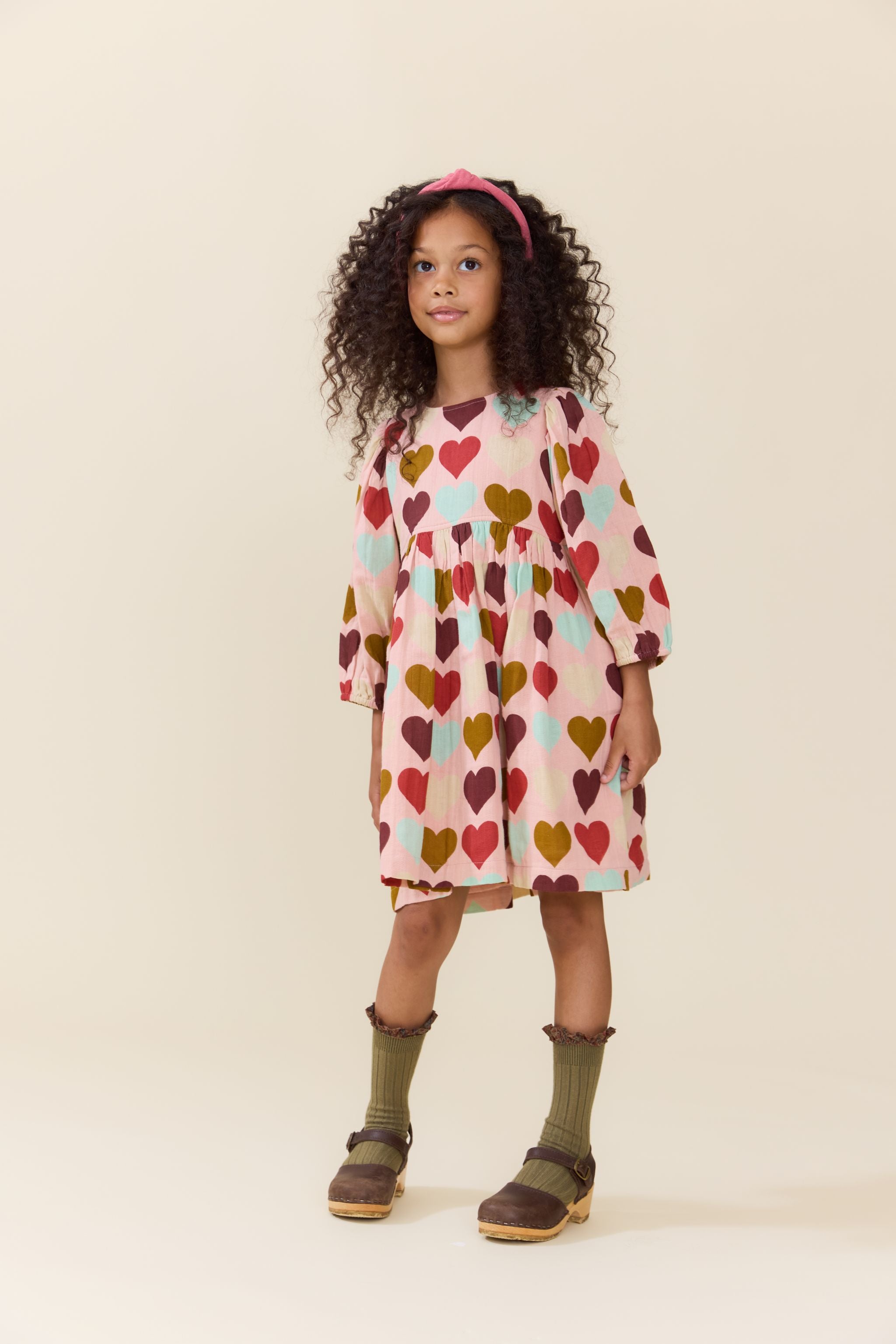 Young girl wearing a Pink Chicken long sleeve dress with colorful harvest heart print styled with a pink headband ribbed socks and brown sandals