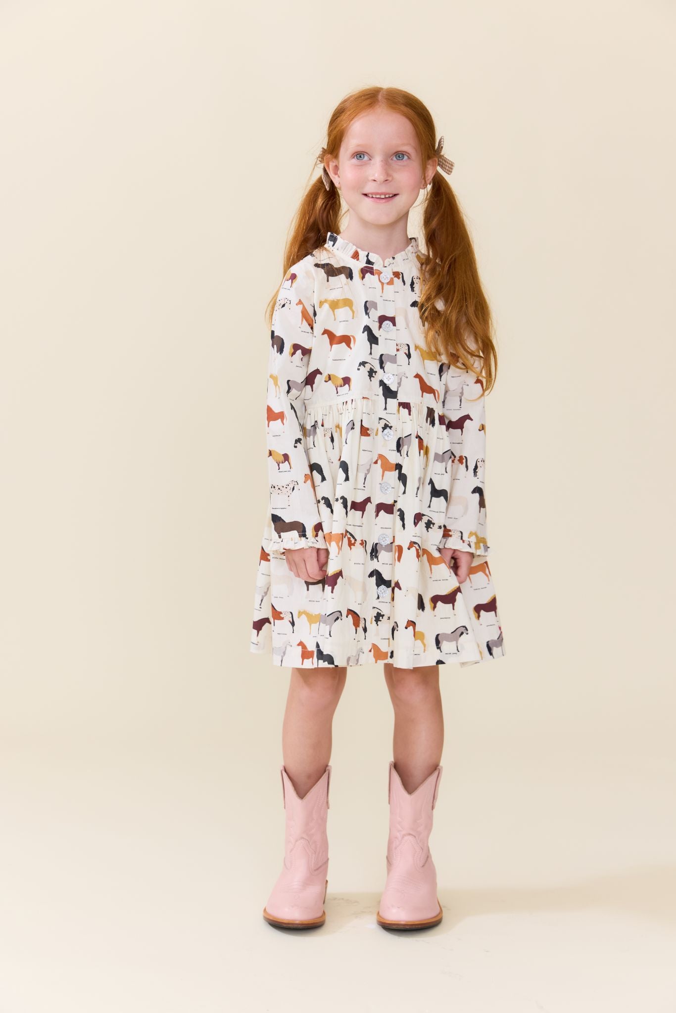 A young girl with red pigtails stands against a solid beige studio background wearing a white long-sleeve dress covered in a colorful horse pattern and light pink cowboy boots.