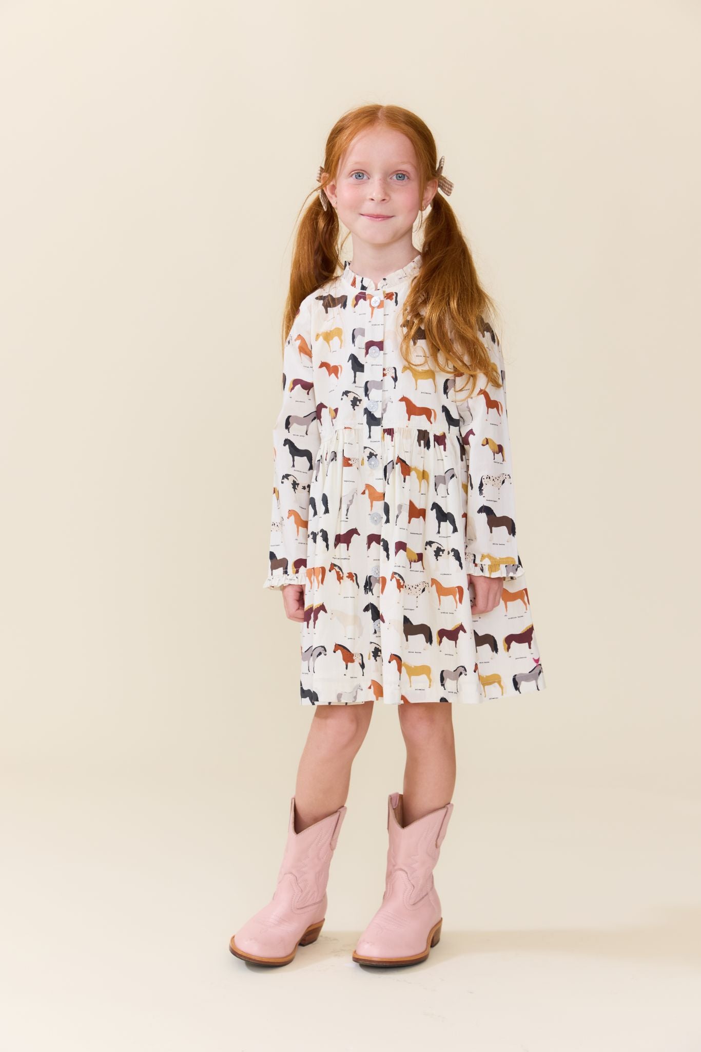 A young girl with red pigtails stands against a solid beige studio background wearing a long-sleeve white dress covered in a colorful horse pattern and light pink cowboy boots.