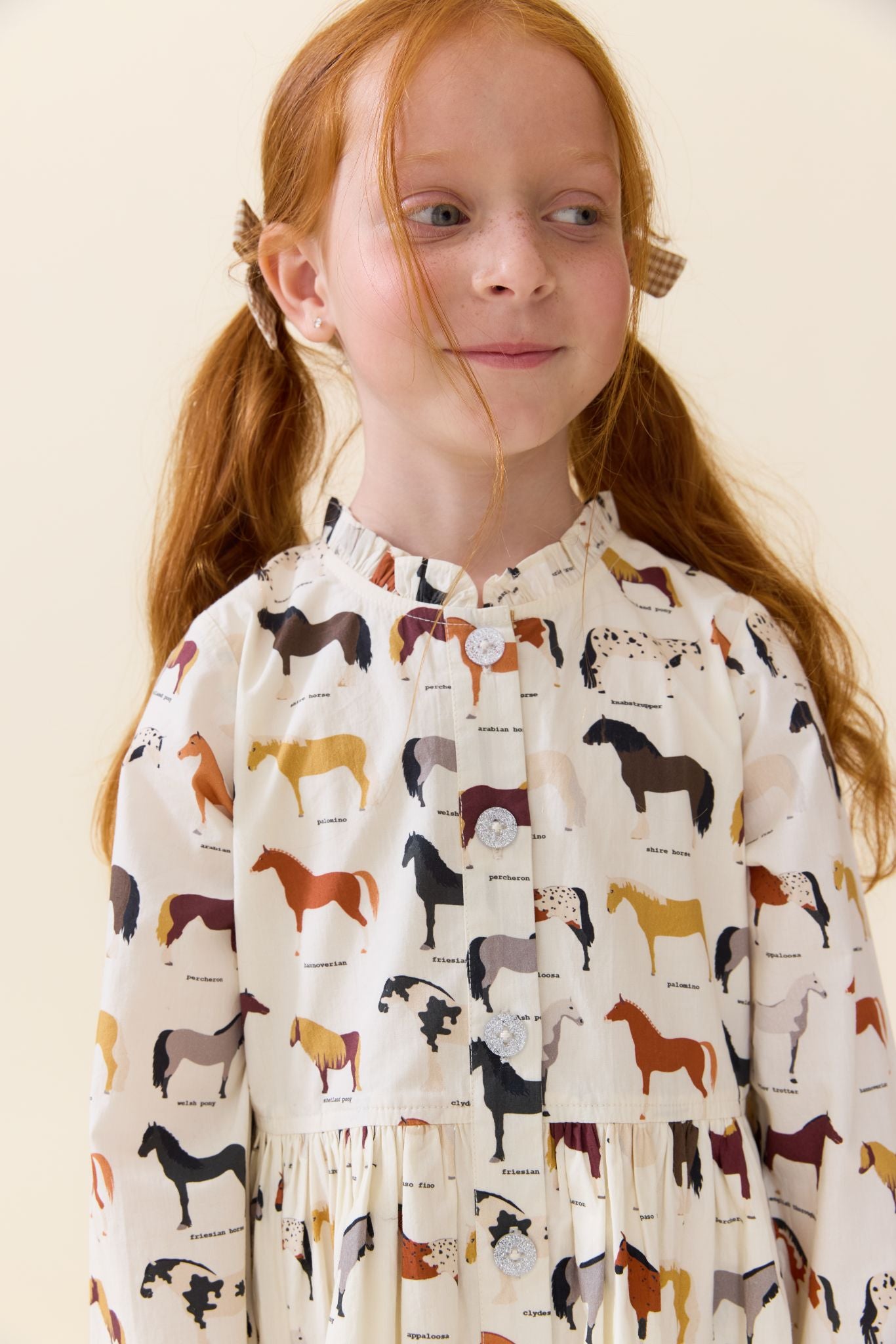 A young girl with red hair in pigtails wearing a cream-colored long-sleeve dress with an all-over horse pattern, photographed against a solid light beige background.