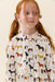 A young girl with red hair in pigtails wearing a cream-colored long-sleeve dress with an all-over horse pattern, photographed against a solid light beige background.