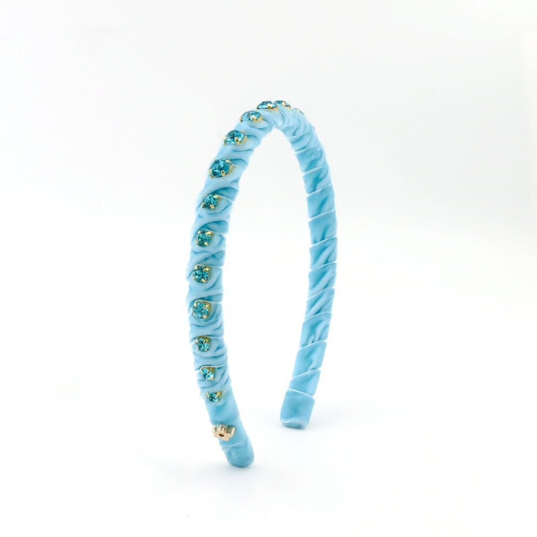 A light blue velvet headband decorated with small blue and green rhinestone clusters and a gold-tone bow at the front, shown on a plain white background.