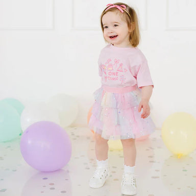 Child wearing a pink shirt and colorful skirt with balloons in the background