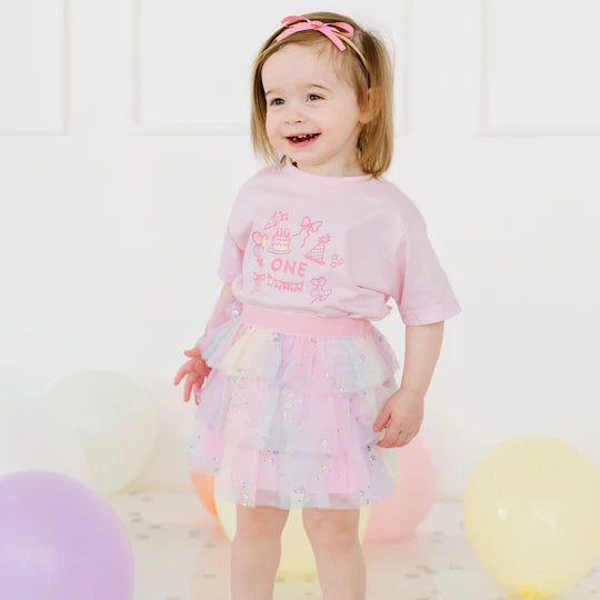 Young girl in a pink outfit with a birthday theme, standing against a white background with balloons.
