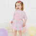 Young girl in a pink outfit with a birthday theme, standing against a white background with balloons.