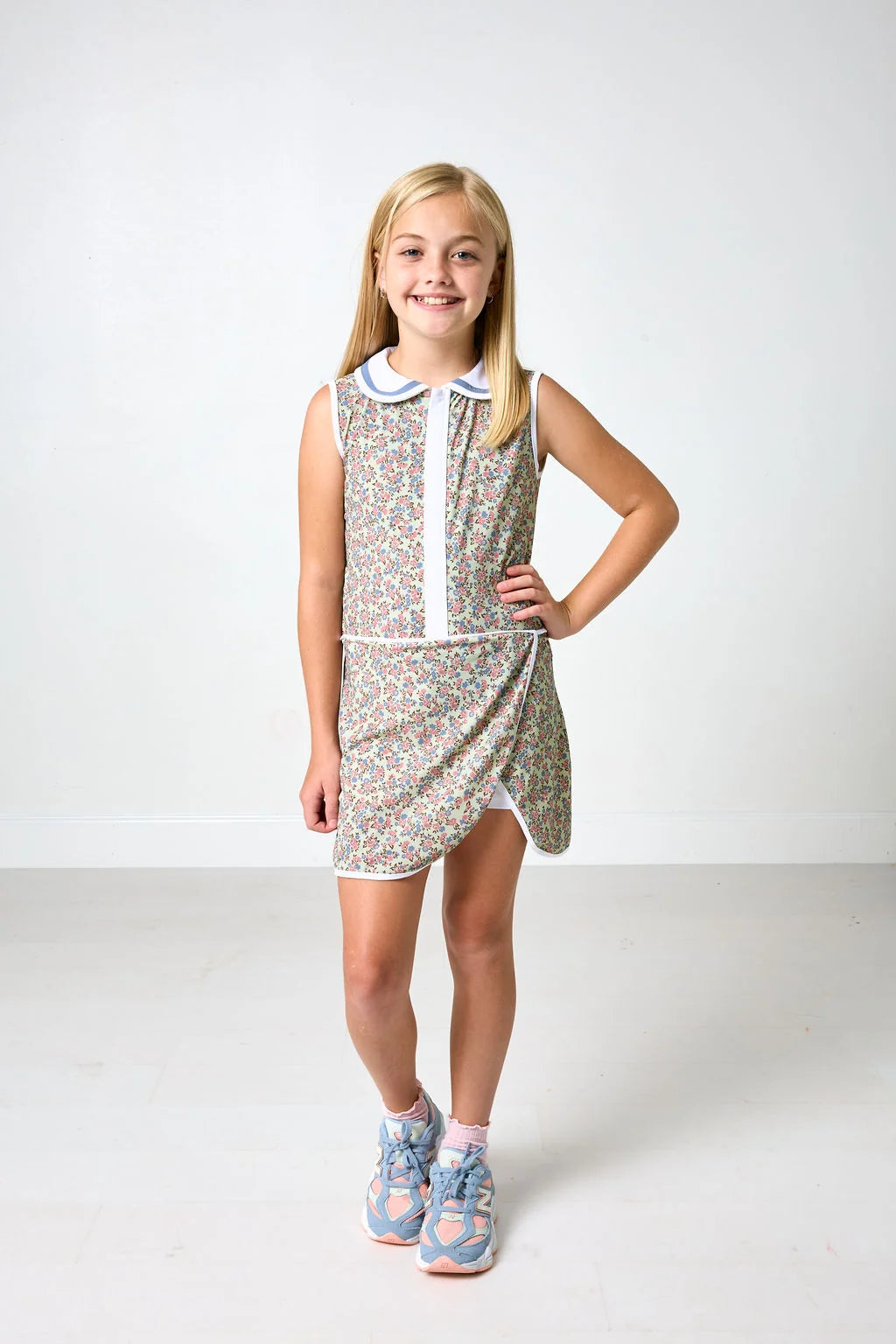 A young girl with blonde hair stands in a studio wearing a sleeveless, patterned mini dress with a white collar and white front placket. She is wearing blue and pink sneakers.