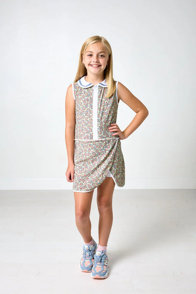 A young girl with blonde hair stands in a studio wearing a sleeveless, patterned mini dress with a white collar and white front placket. She is wearing blue and pink sneakers.