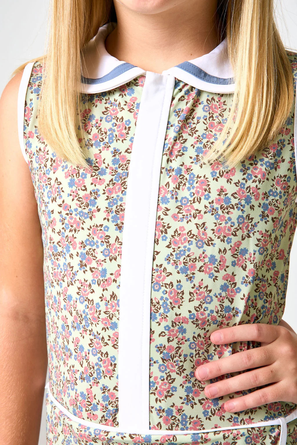 Close-up of a sleeveless floral dress with a white collar trimmed in blue, featuring a white front placket and a pink, blue, and green floral pattern on a light beige background.