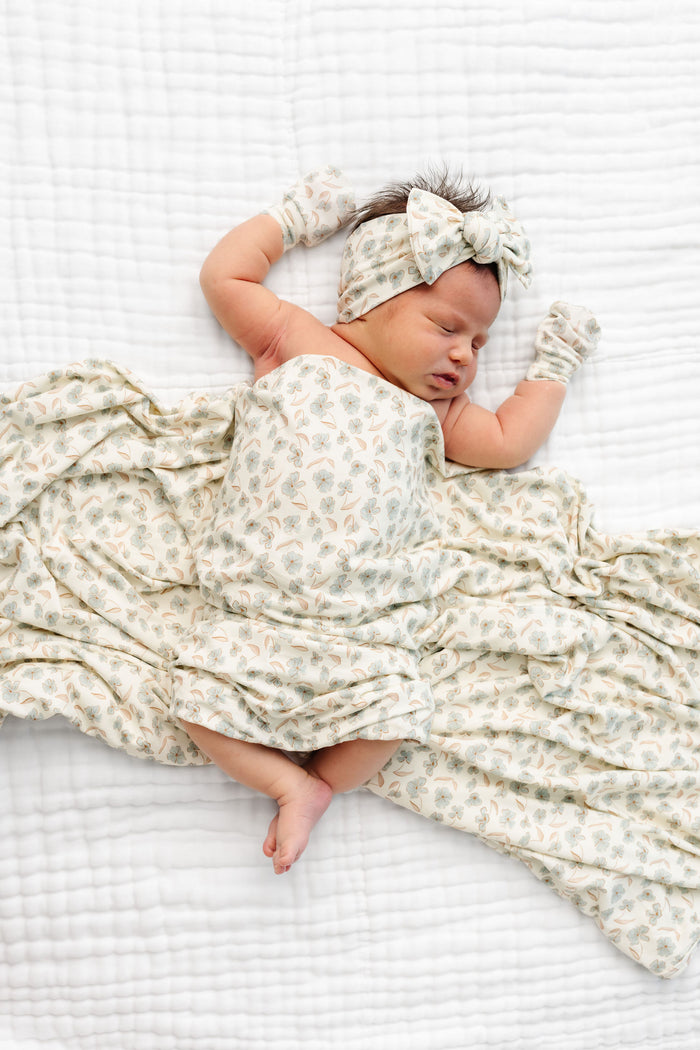 A baby lying in blanet with a delicate floral print, a matching bow headband, and two matching mittens, all in a soft cream color with light blue and tan floral motifs.