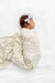 A newborn baby lying on a white textured blanket, swaddled in a cream-colored swaddle blanket with a delicate green and beige floral pattern, and wearing a matching light blue bow headband.