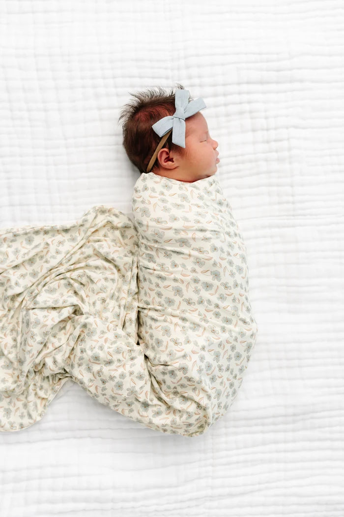 A newborn baby lying on a white textured blanket, swaddled in a cream-colored swaddle blanket with a delicate green and beige floral pattern, and wearing a matching light blue bow headband.
