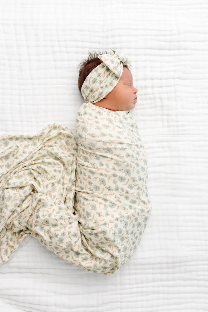 A newborn baby wrapped in a soft patterned swaddle blanket and matching headband, lying on a white textured muslin blanket.