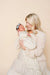 A blonde woman in a cream dress holds a sleeping newborn baby wrapped in a cream and sage green floral patterned swaddle blanket. The baby also wears a light blue bow headband.