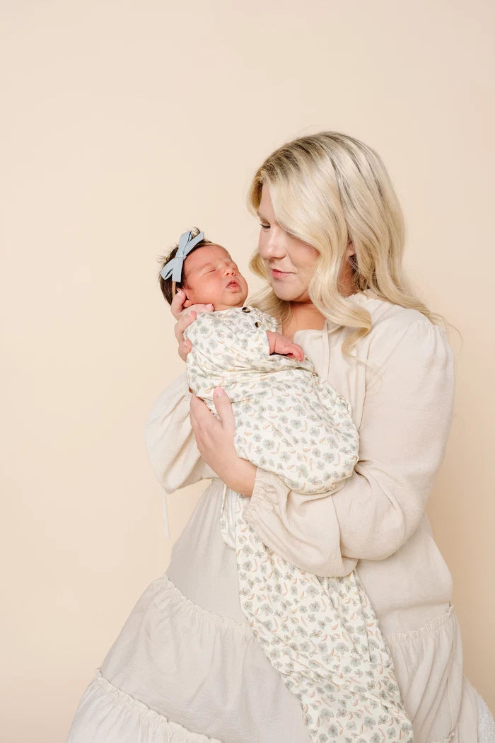 A blonde woman in a cream dress holds a sleeping newborn baby wrapped in a cream and sage green floral patterned swaddle blanket. The baby also wears a light blue bow headband.
