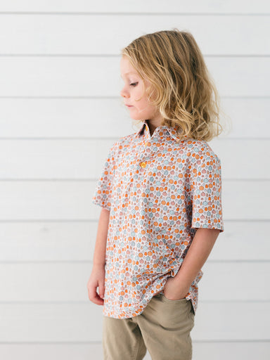 A young child wearing a short-sleeve button-up shirt with an orange, white, and gray pumpkin pattern, paired with khaki pants, standing against a white paneled wall.