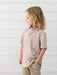 A young child wearing a short-sleeve button-up shirt with an orange, white, and gray pumpkin pattern, paired with khaki pants, standing against a white paneled wall.