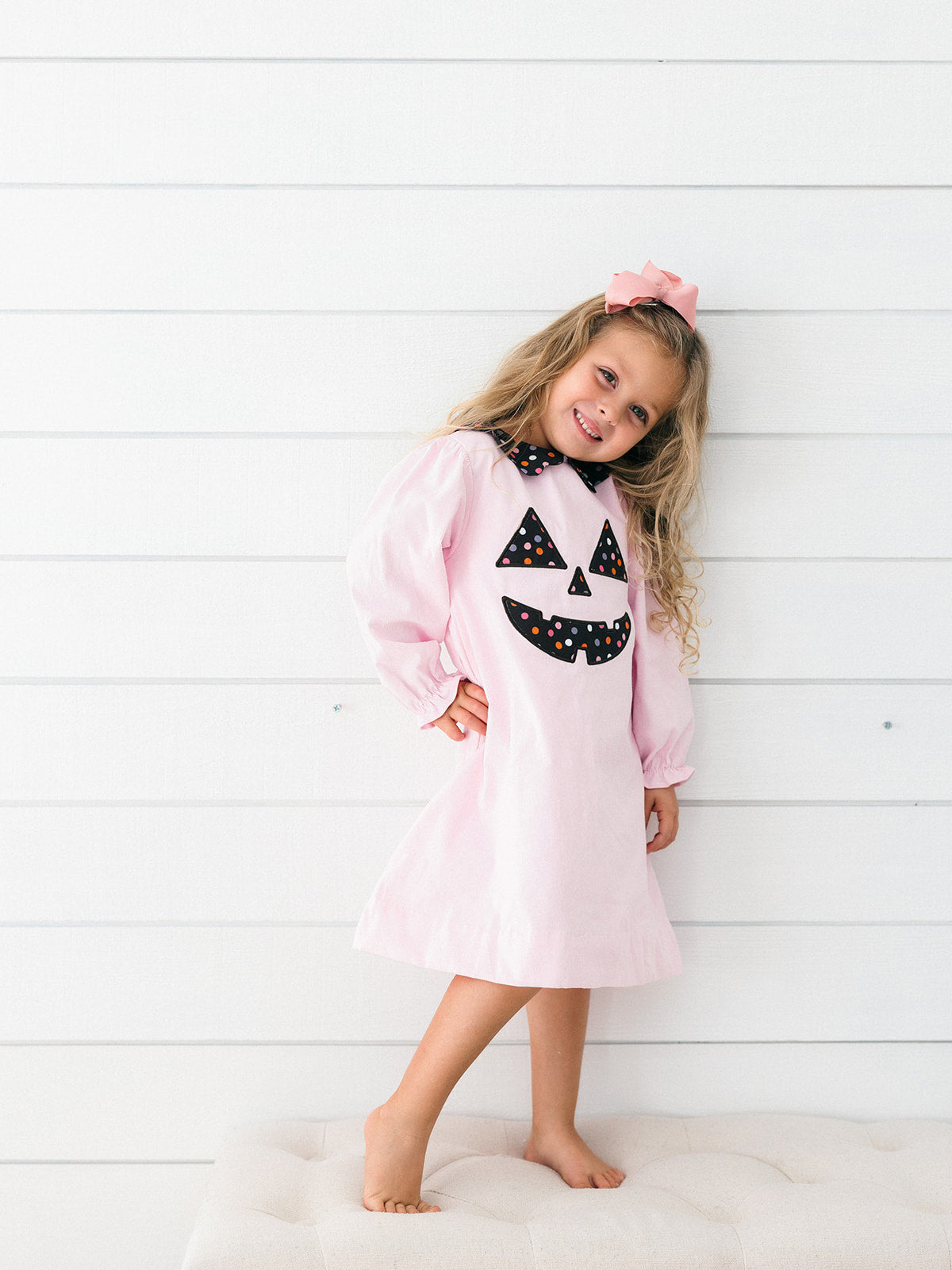 A young girl with blonde hair wearing a light pink long-sleeve dress with a black and white polka-dot jack-o'-lantern face on the front, standing barefoot against a white shiplap wall.
