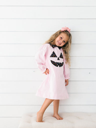A young girl with blonde hair wearing a light pink long-sleeve dress with a black and white polka-dot jack-o'-lantern face on the front, standing barefoot against a white shiplap wall.