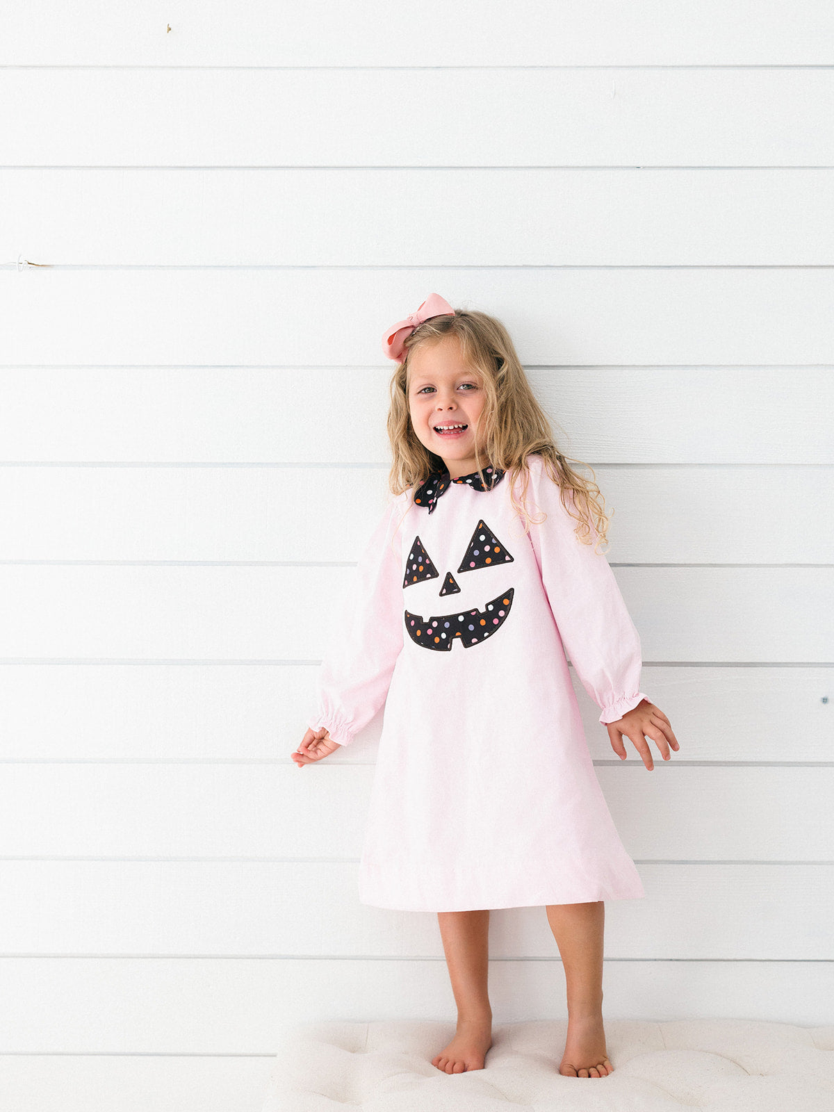 A young girl with blonde hair wearing a light pink long-sleeve nightgown with a black and white polka-dot jack-o'-lantern face applique, standing against a white wood-paneled wall.