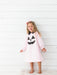 A young girl with blonde hair wearing a light pink long-sleeve nightgown with a black and white polka-dot jack-o'-lantern face applique, standing against a white wood-paneled wall.