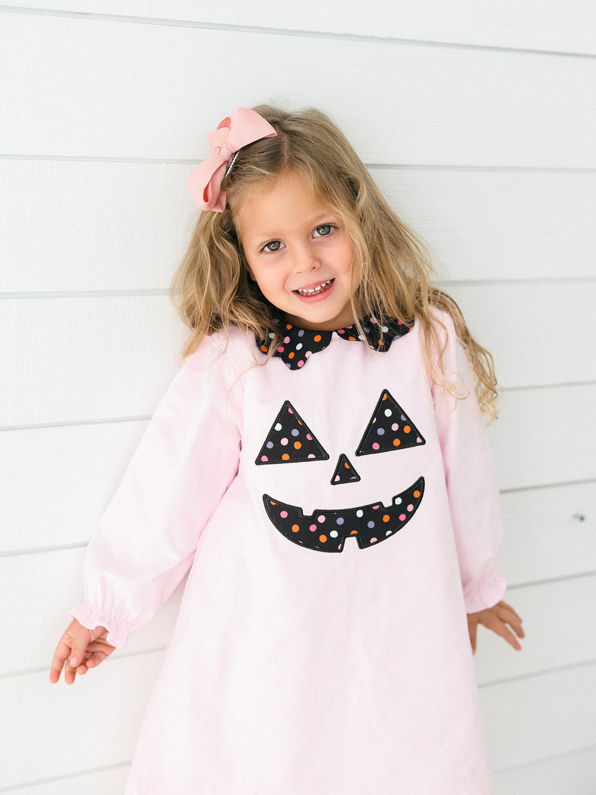 A young girl with blonde hair and a pink bow stands against a white paneled wall wearing a light pink long-sleeve dress with a black polka-dot jack-o'-lantern face and bow collar.