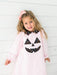 A young girl with blonde hair and a pink bow stands against a white paneled wall wearing a light pink long-sleeve dress with a black polka-dot jack-o'-lantern face and bow collar.