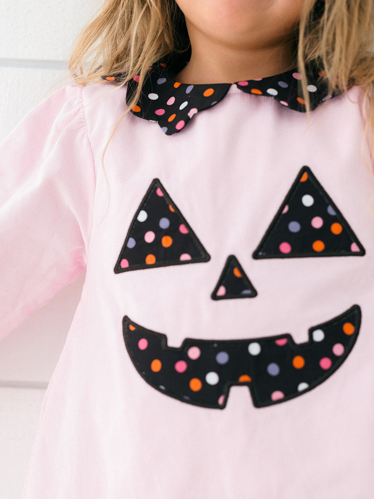 A close-up of a child wearing a light pink long-sleeve dress with a black Peter Pan collar and a black jack-o'-lantern face applique decorated with multicolored polka dots.