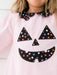 A close-up of a child wearing a light pink long-sleeve dress with a black Peter Pan collar and a black jack-o'-lantern face applique decorated with multicolored polka dots.