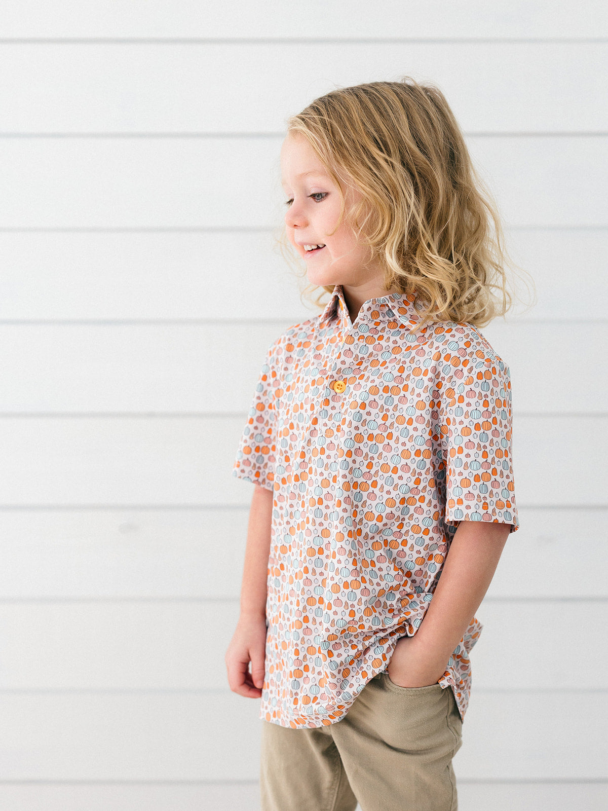 A young girl with blonde hair is shown in profile wearing a short-sleeve button-up shirt with an orange, white, and light blue pumpkin pattern, paired with khaki pants, set against a white shiplap wall.