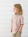 A young girl with blonde hair is shown in profile wearing a short-sleeve button-up shirt with an orange, white, and light blue pumpkin pattern, paired with khaki pants, set against a white shiplap wall.