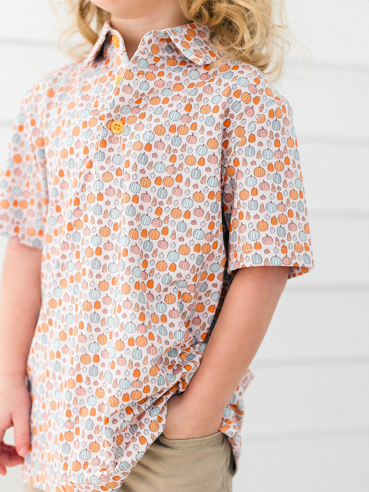 A child wearing a short-sleeve button-up shirt with an all-over pumpkin print in orange, gray, and white, paired with khaki pants against a white paneled wall.