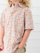 A child wearing a short-sleeve button-up shirt with an all-over pumpkin print in orange, gray, and white, paired with khaki pants against a white paneled wall.