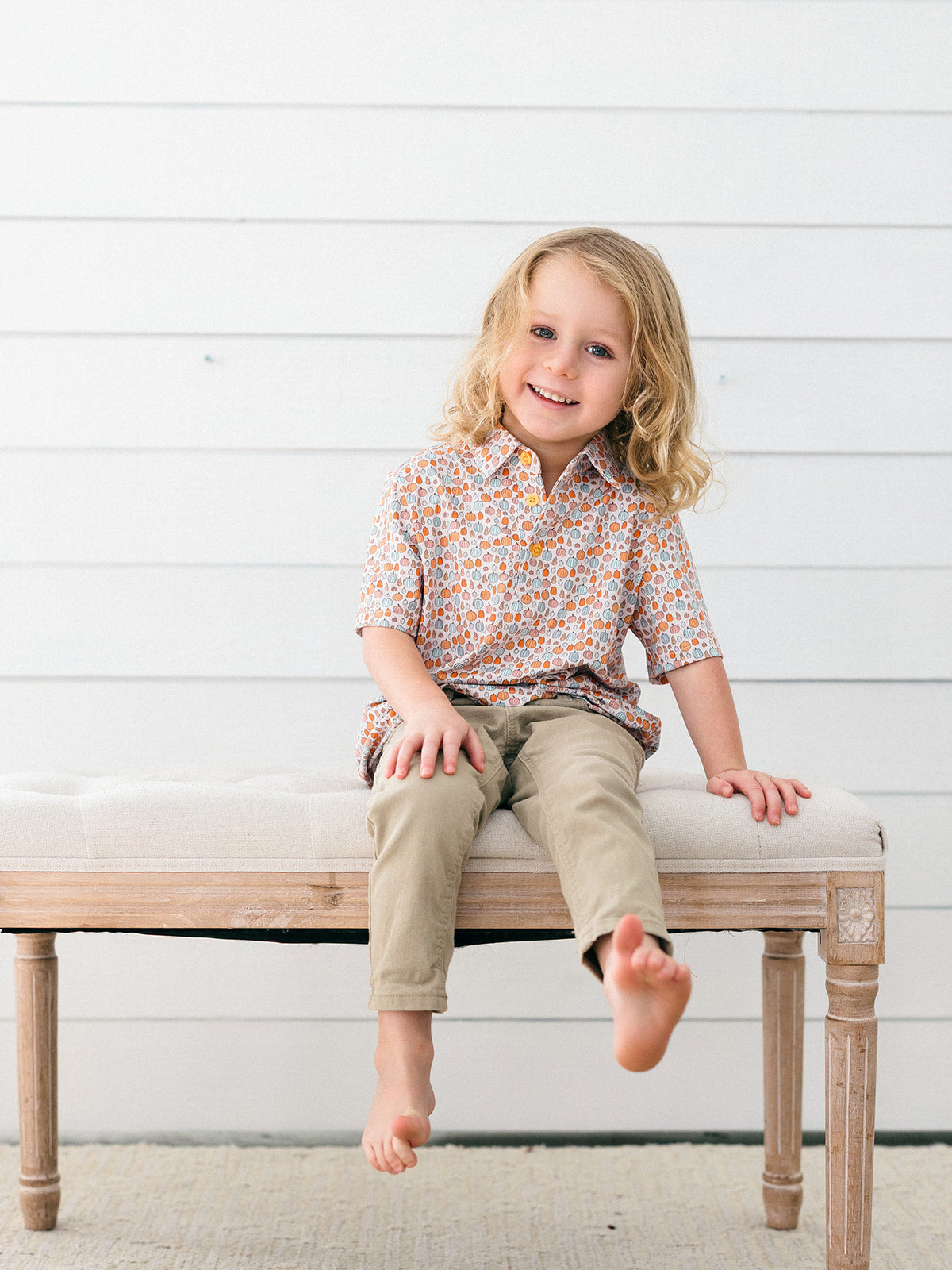 A young child with blonde hair sits barefoot on a light wood bench with a cream cushion, wearing a short-sleeve button-up shirt with a small orange, beige, and white pumpkins, and khaki pants, against a white shiplap wall.