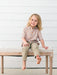 A young child with blonde hair sits barefoot on a light wood bench with a cream cushion, wearing a short-sleeve button-up shirt with a small orange, beige, and white pumpkins, and khaki pants, against a white shiplap wall.