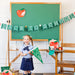 Young girl in a school uniform standing in front of a green chalkboard with 'Back to School' decorations.