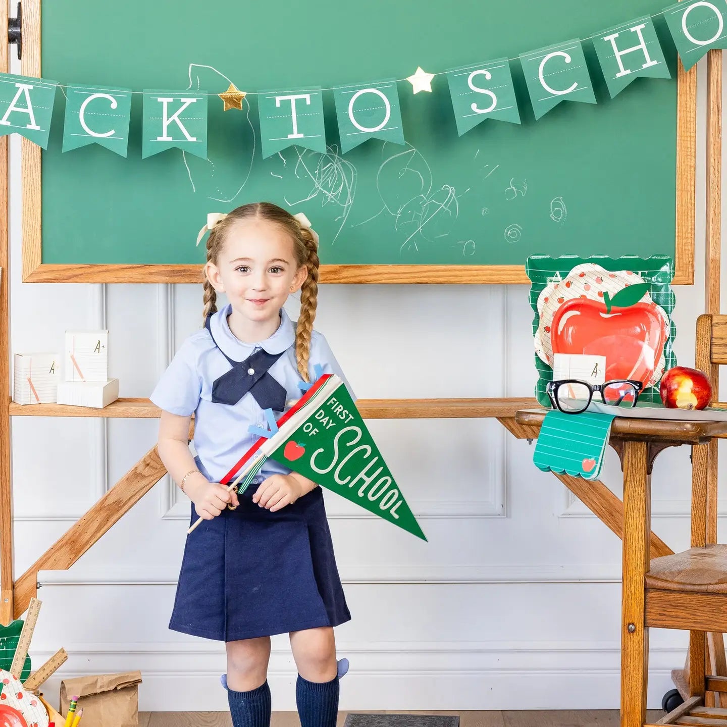 Young girl in a school uniform holding a 'First Day of School' flag in front of a green chalkboard with 'Back to School' decorations.