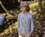 A young boy wearing a heather gray crewneck sweatshirt featuring a vintage-style graphic of a fisherman holding a fishing rod with the words 'FISH WEARABLE' above a large fish. A small black brand tag is visible on the lower right hem.