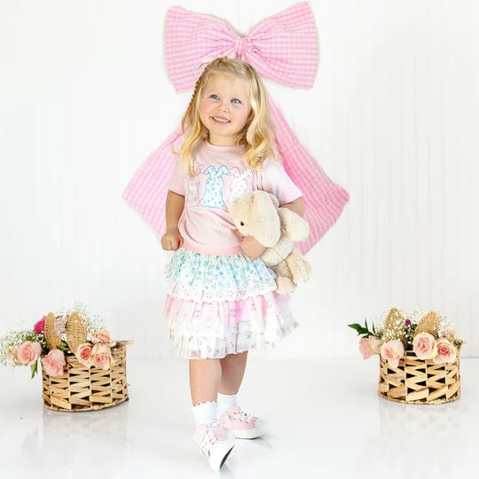 Young girl in a pink outfit with a large bow, holding a teddy bear, on a white background.