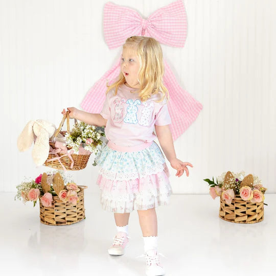 Young girl in a pastel outfit with a large pink bow, holding a basket with flowers and a toy rabbit.