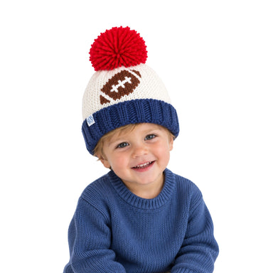 A smiling toddler wearing a navy blue knit beanie with a red pom-pom and a brown football motif on the side, paired with a blue knit sweater against a plain white background.
