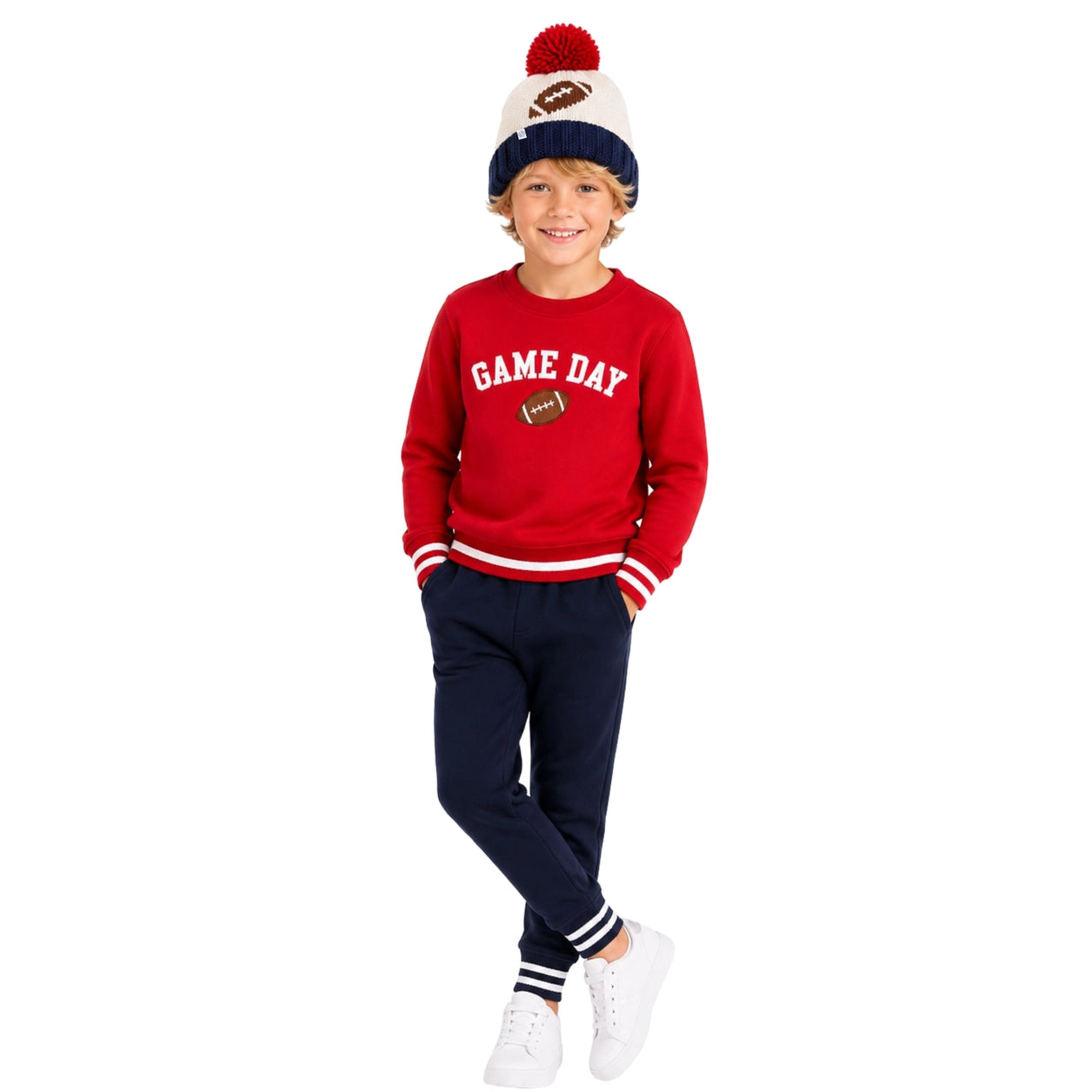 A young boy modeling a red crewneck sweatshirt with 'GAME DAY' in white varsity lettering and a football graphic, paired with navy sweatpants, white sneakers, and a cream and navy beanie with a red pom-pom.
