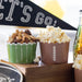 Two snack cups with popcorn and nuts on a wooden tray, with a 'Let's Go!' banner in the background.