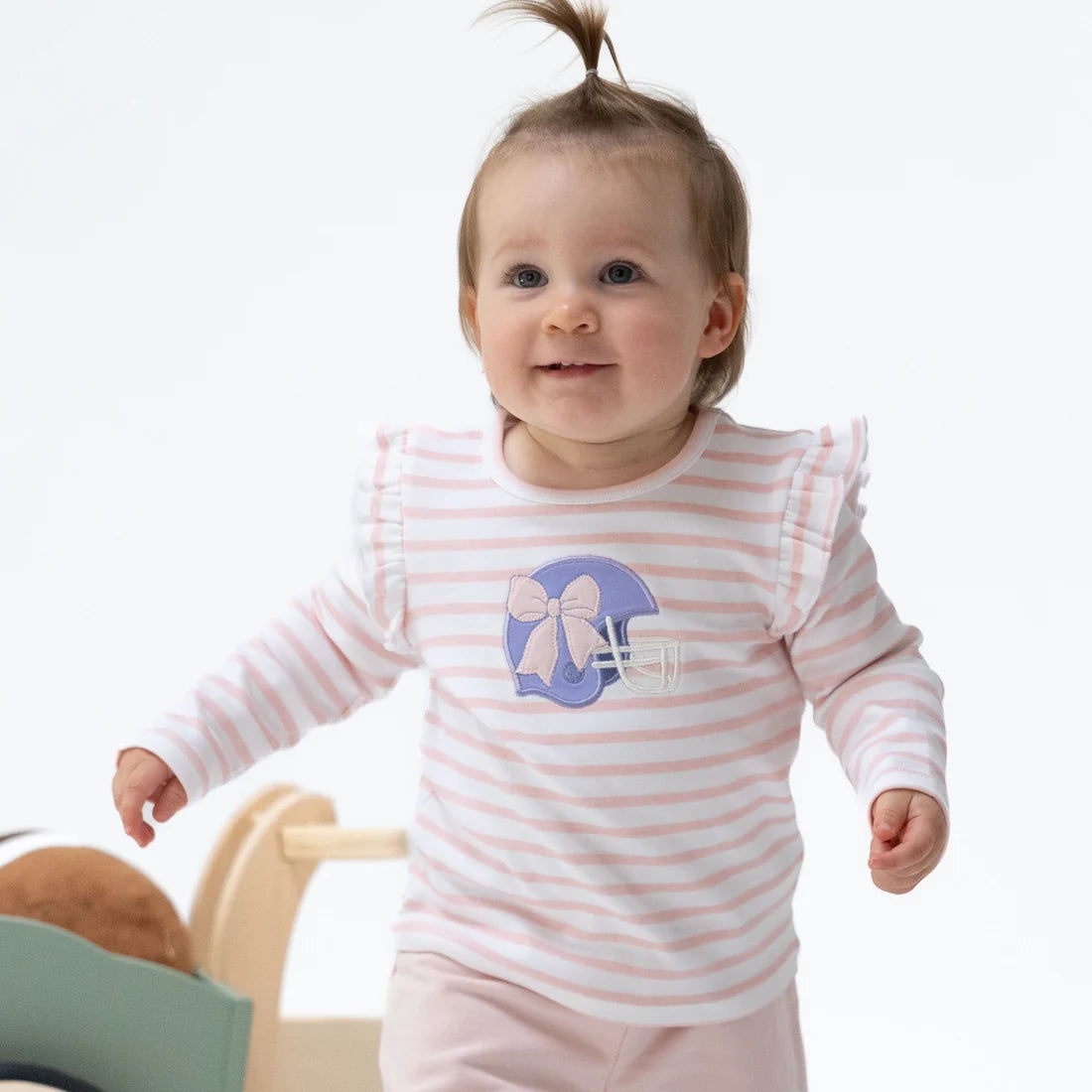 A baby girl wearing a light pink and white striped long-sleeve shirt with a purple football helmet graphic on the chest, standing against a clean white background.