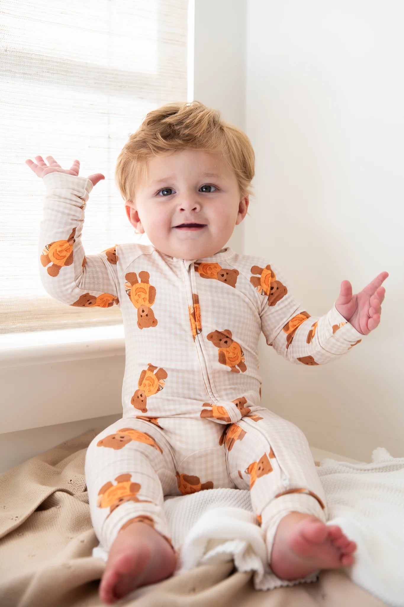 A baby sitting on a bed wearing a cream-colored waffle-knit footed pajama suit with an orange teddy bear pattern, with arms raised in excitement.