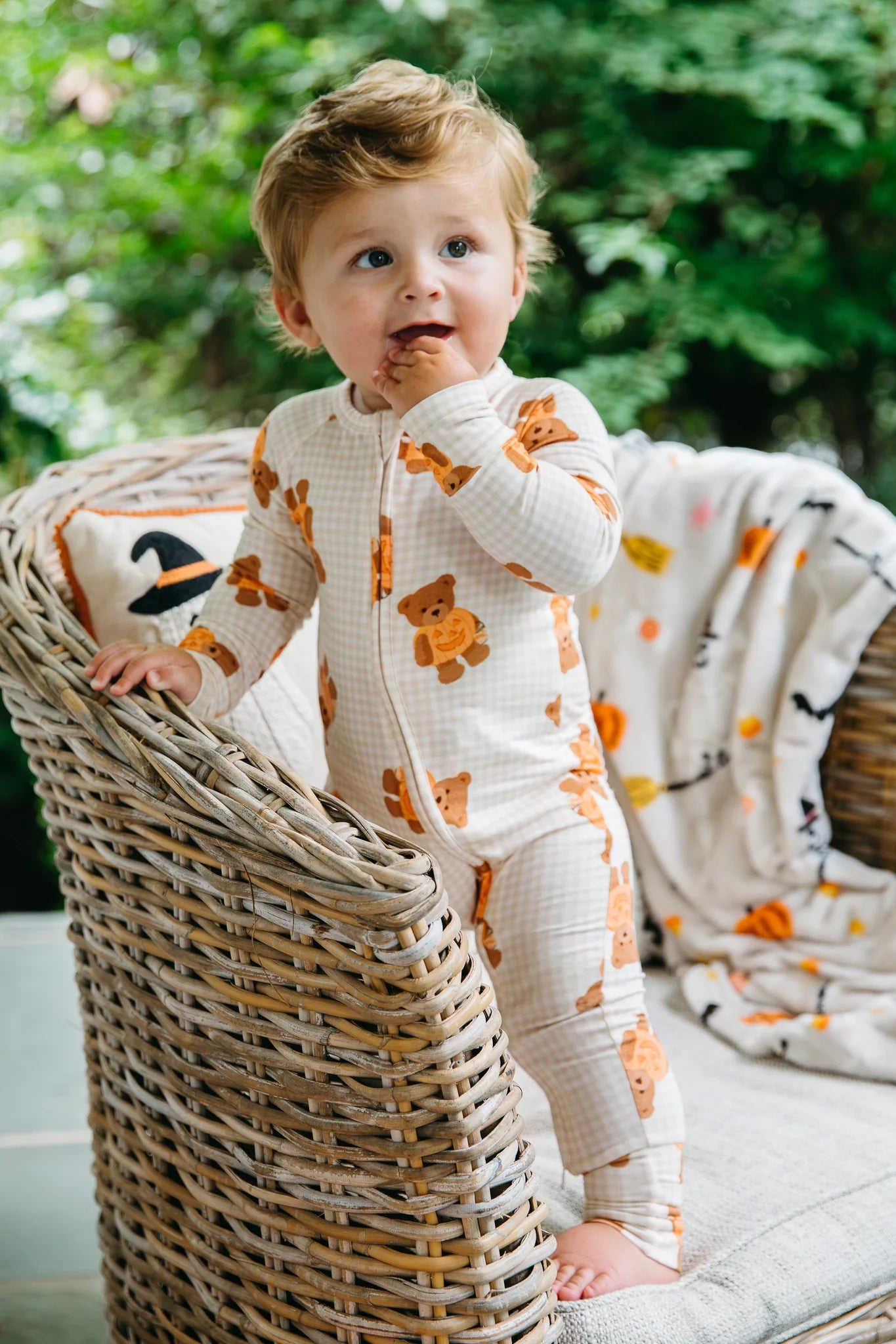 A toddler stands on a wicker chair wearing a cream-colored gingham one-piece pajama with orange teddy bear prints, with a matching patterned throw blanket draped over the chair in an outdoor garden setting.
