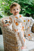 A toddler stands on a wicker chair wearing a cream-colored gingham one-piece pajama with orange teddy bear prints, with a matching patterned throw blanket draped over the chair in an outdoor garden setting.