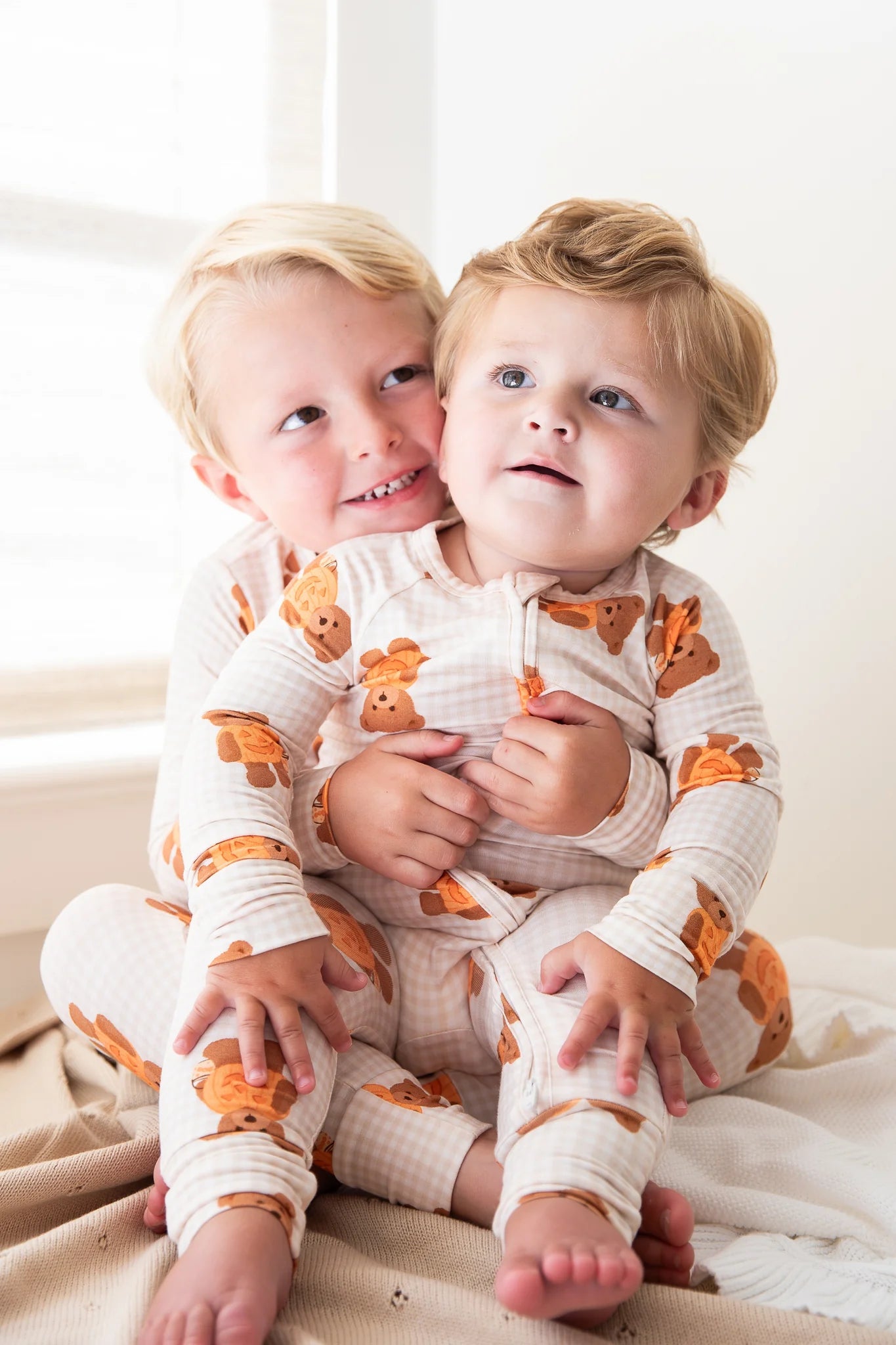 Two young children sitting together indoors, wearing matching long-sleeve footless pajamas with an orange teddy bear print on a cream background.